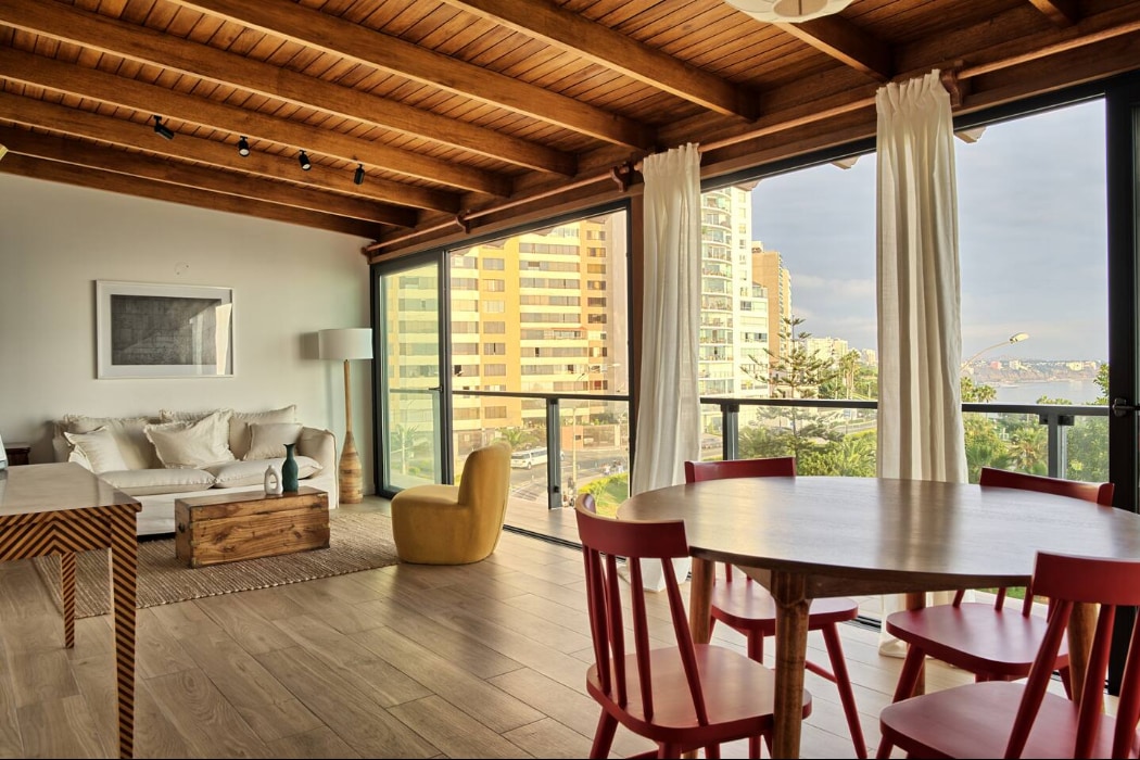 A spacious living area is highlighted by large glass windows that offer a view of the surroundings. A comfortable sofa and a light wood coffee table are present, complemented by a yellow accent chair and a round dining table with red chairs. Wooden beams adorn the ceiling.