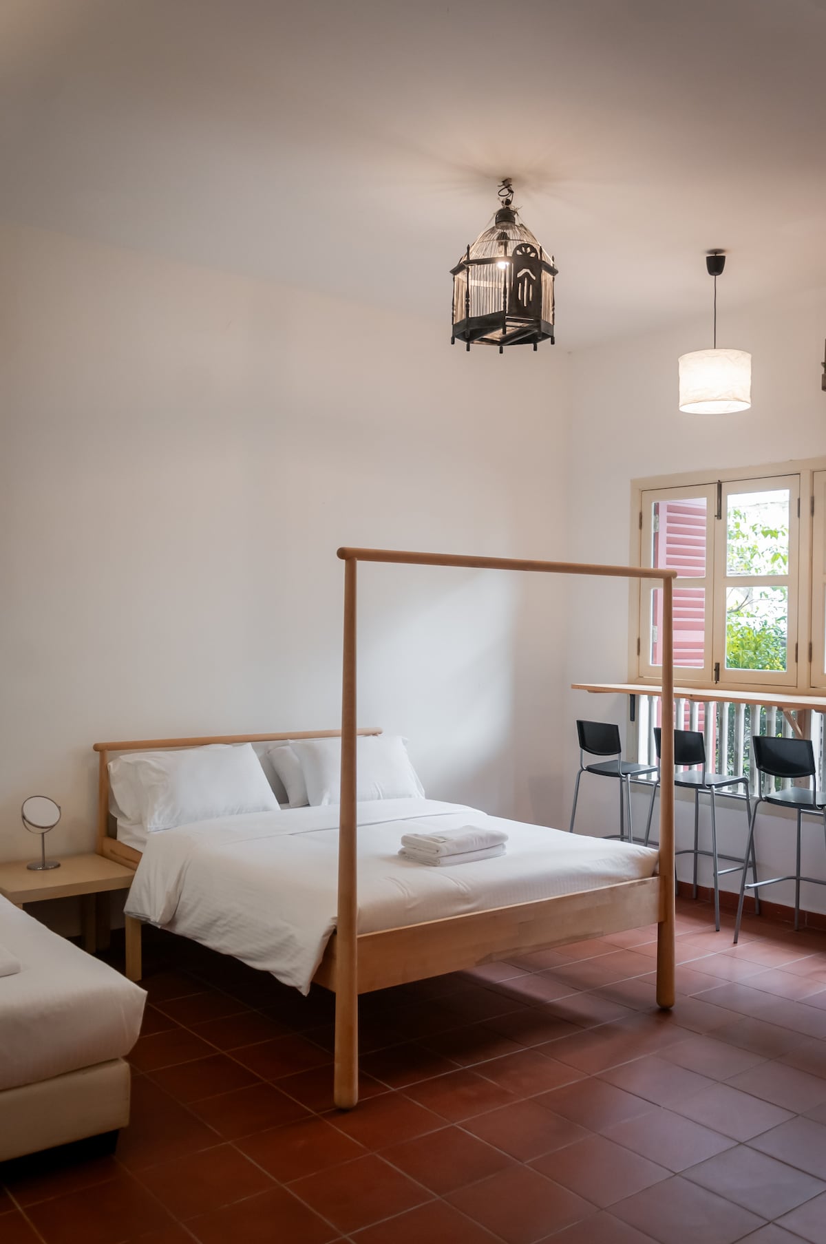 A minimalistic room features a queen-size bed with crisp linens and a cozy single bed nearby. Natural light filters through traditional wooden windows, complementing the simple furnishings, which include a small high bar table and modern chairs.