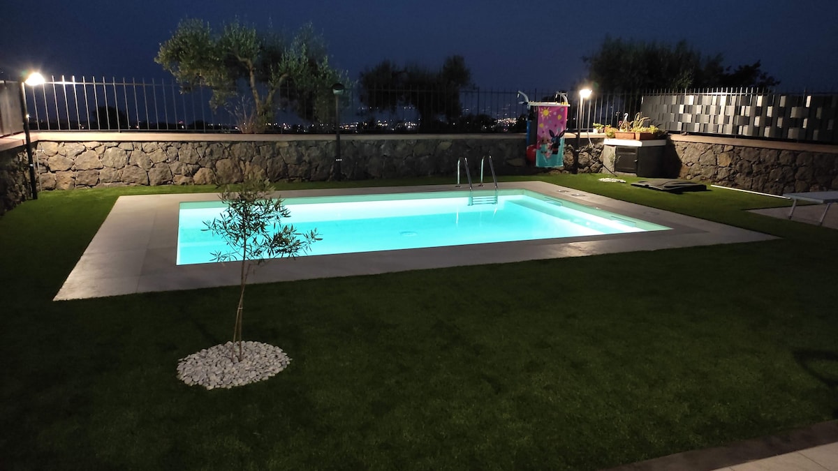 The illuminated swimming pool is surrounded by well-kept grass and features a smooth, light-colored deck. Soft lighting casts a gentle glow on the pool, enhancing the tranquil evening atmosphere. The background showcases a garden area with low stone walls and distant views of the landscape.