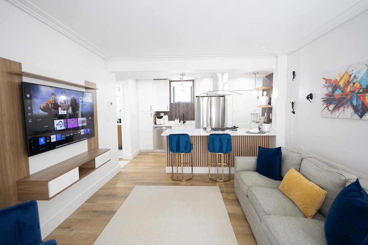 A modern living area is displayed, featuring a stylish gray sofa adorned with a yellow accent pillow. A mounted television offers entertainment options, while a light-colored area rug adds warmth. The open layout connects to a contemporary kitchen with sleek finishes and a neutral color palette.