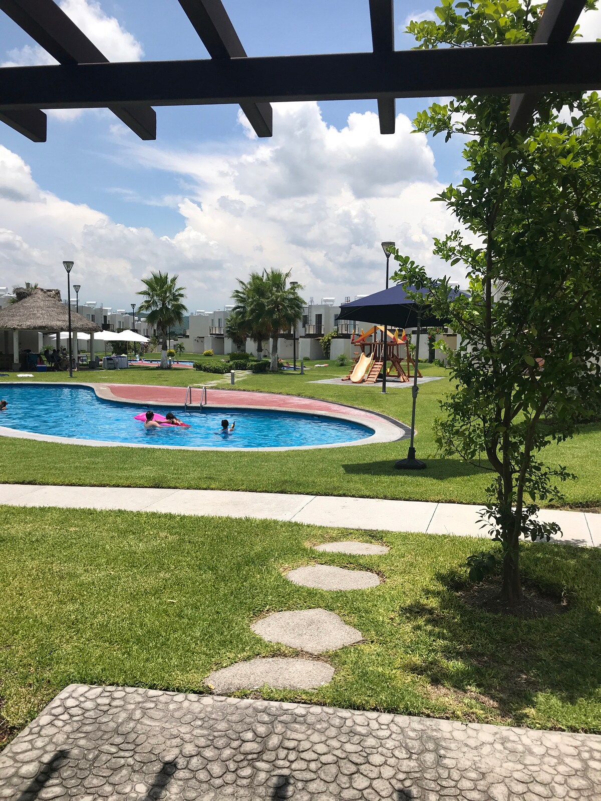 A clear view of a pool surrounded by lush greenery, where children can be seen playing. The pool area is complemented by shaded seating and a nearby playground, creating a family-friendly environment. Pathways meander through the grass, inviting exploration.