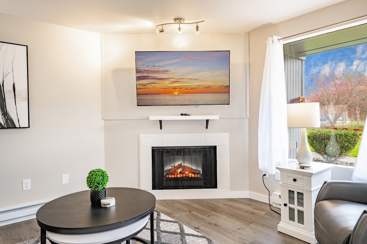 A cozy living area is highlighted by a modern electric fireplace and a wall-mounted TV displaying a scenic sunset. Natural light enters through a nearby window, complemented by soft white curtains. A round coffee table sits at the center, enhancing the inviting space.