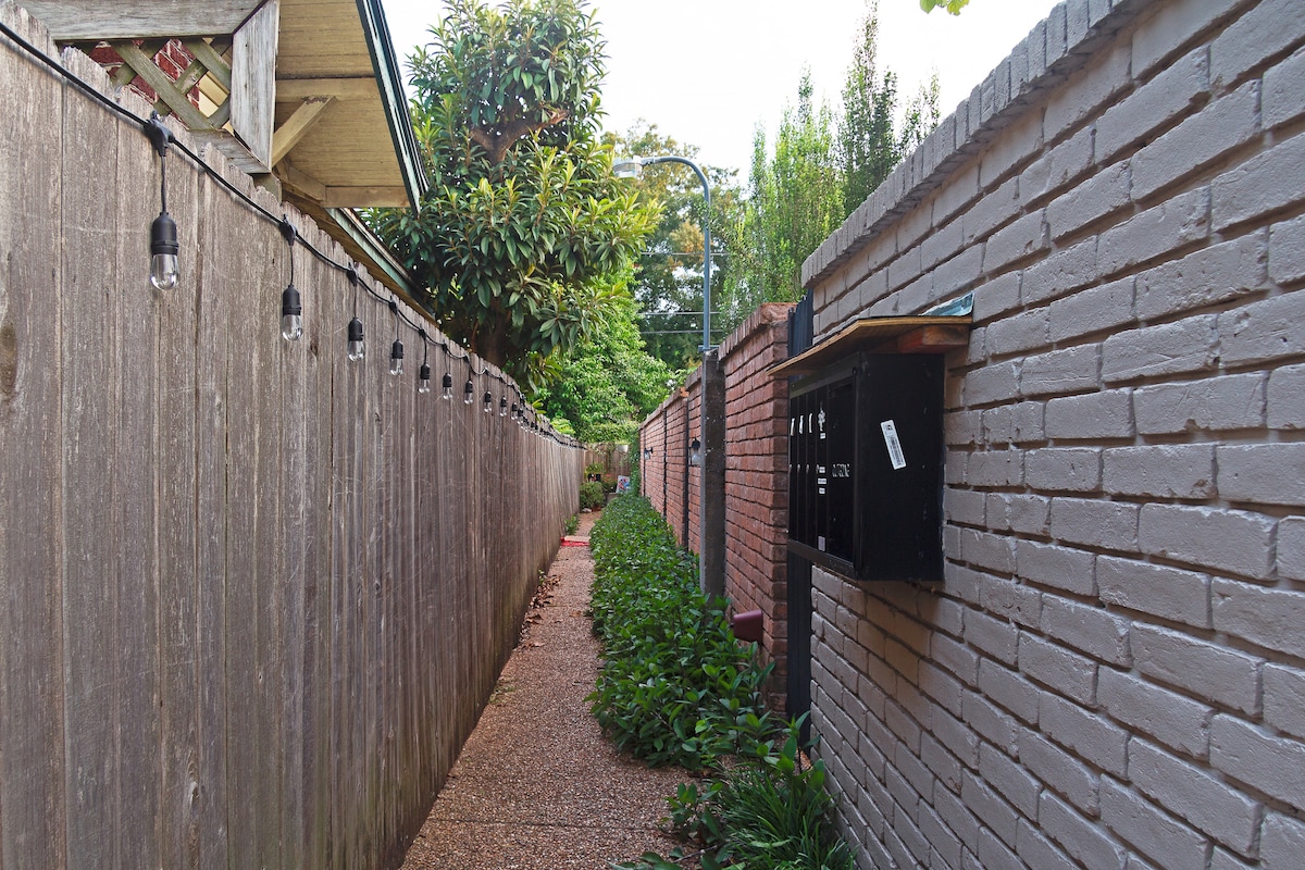 A narrow pathway is lined with wooden fences and brick walls in a serene environment. Lush greenery and small plants border the gravel path. Mailboxes are positioned on the right side, providing functional details to the charming setting.