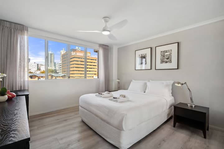 Elegantna spavaća soba s ventilatorom, velikim prozorom i mekim dekorom s pogledom na grad.