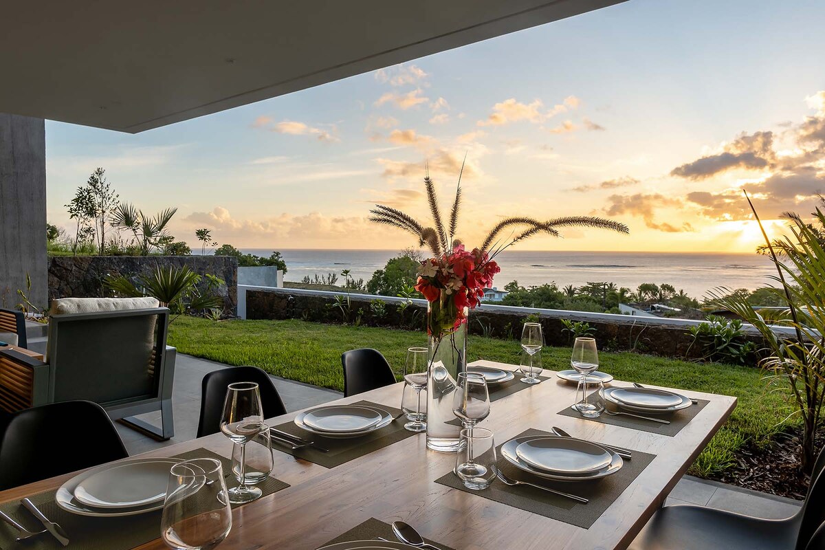A spacious dining area is set with a wooden table, complemented by elegant glassware and tableware. A vase containing fresh flowers adds a natural touch. The backdrop features a scenic sunset over the ocean and lush greenery, creating a serene outdoor dining experience.