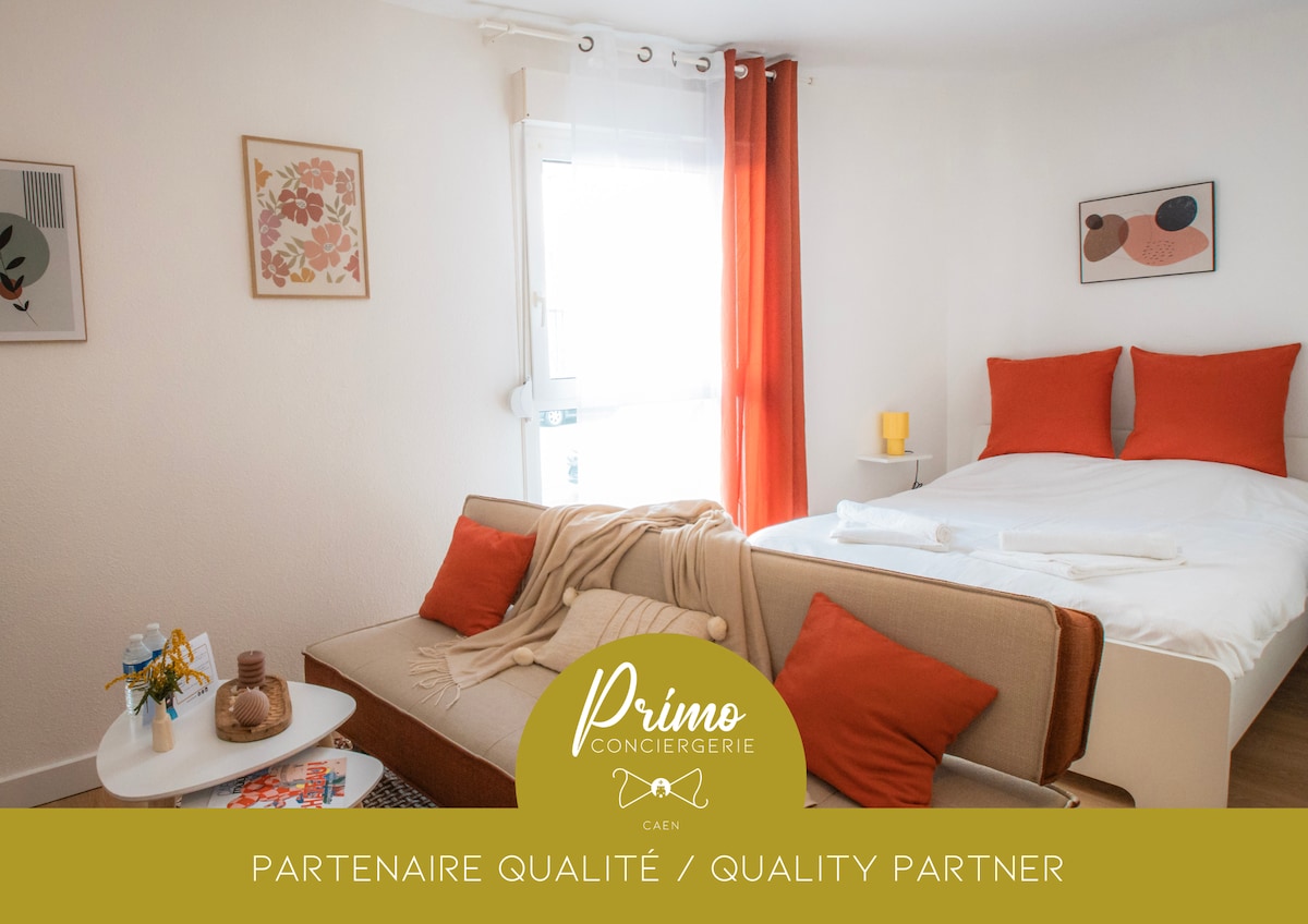 A bright studio features a spacious bed with vibrant orange pillows, complemented by a cozy sofa adorned with similar colored cushions. Natural light filters through sheer curtains, creating a warm ambiance. A small table with refreshments is visible, enhancing convenience for guests.
