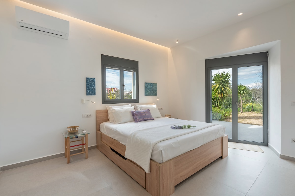 A bright bedroom features a spacious bed with soft linens and a decorative pillow. Large glass doors open to a private outdoor area, revealing greenery outside. Wall-mounted lights provide gentle illumination, and artwork hangs on either side of the windows, enhancing the serene atmosphere.