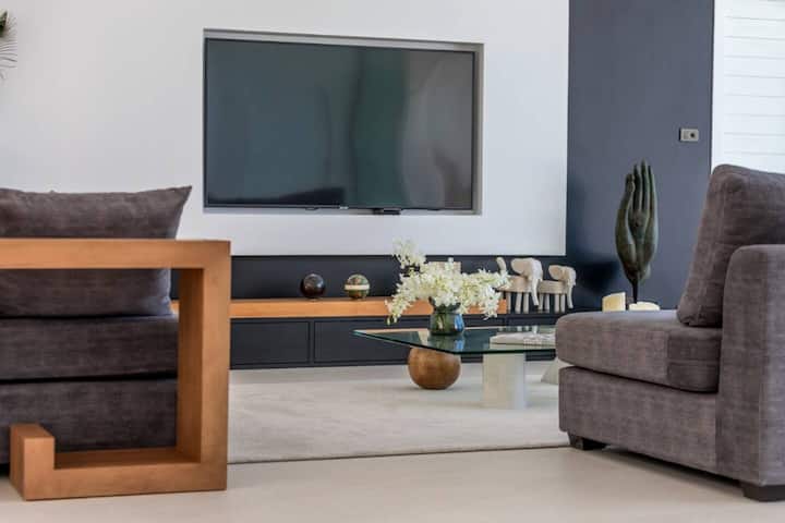 Sala de estar contemporánea con elegantes sofás grises, una mesa de café de vidrio con una base redondeada y un televisor de pantalla plana montado en la pared. La decoración incluye una escultura de mano moderna, figuras de elefante y flores blancas frescas, creando un ambiente sofisticado.