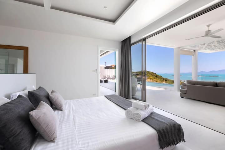Despiértate con impresionantes vistas al mar desde este moderno dormitorio con una lujosa cama tamaño queen con detalles en gris. Las puertas de vidrio del piso al techo se abren a una amplia terraza con una piscina infinita, tumbonas y una acogedora zona de estar al aire libre.