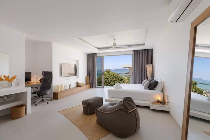 Amplio dormitorio con una lujosa cama tamaño queen, cómodos sillones y un espacio de trabajo dedicado. Las puertas de vidrio del piso al techo se abren a un balcón privado con vistas panorámicas al mar. La decoración moderna y la luz natural crean un refugio sereno y elegante.