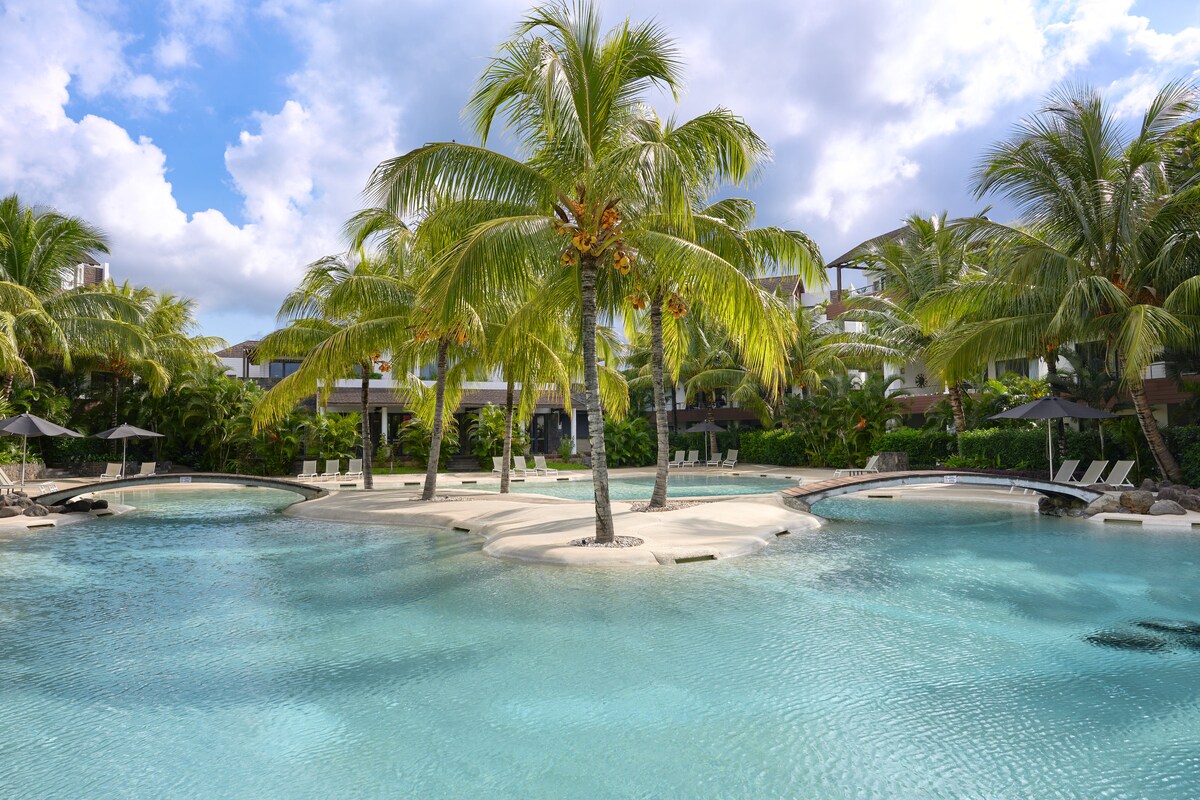 A tranquil lagoon-style pool is surrounded by lush palm trees, providing a refreshing oasis. Sun loungers are positioned on the sandy edges, inviting relaxation. The clear blue water reflects the sky, enhancing the serene atmosphere of the outdoor space.