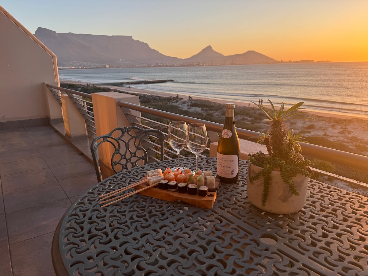 An outdoor dining table is set on a private balcony, displaying a platter of sushi, a bottle of sparkling wine, and two glasses. Scenic views of the ocean and sunset can be seen in the background, with Table Mountain and coastal features enhancing the serene atmosphere.