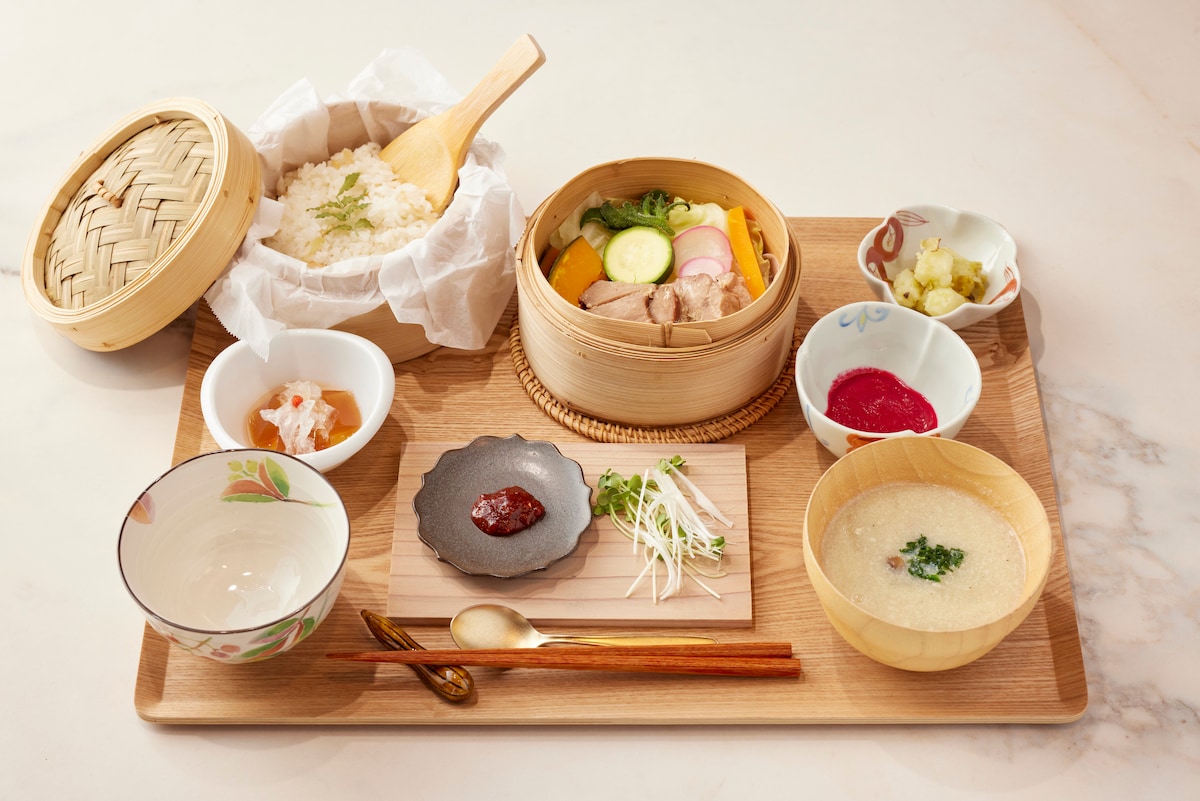 A wooden serving tray presents a well-arranged traditional meal. Steamed rice in a bamboo container is accompanied by a wooden bowl of creamy soup, colorful vegetables in a steaming basket, and various small dishes featuring pickled vegetables, sauce, and a delicate jelly. A spoon and chopsticks rest alongside.