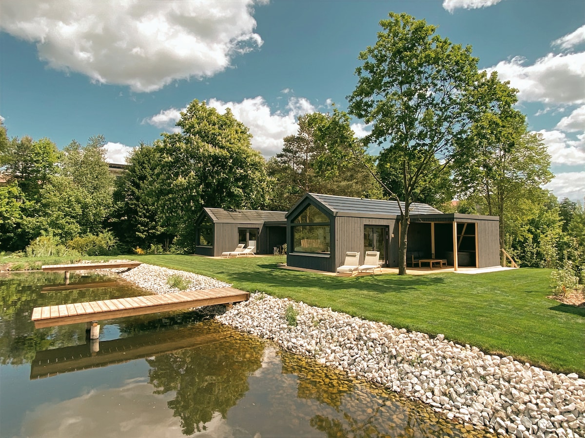 Two modern cabins are situated amidst lush greenery, reflecting in a tranquil body of water. A wooden deck extends into the water, lined with smooth stones. Comfortable lounge chairs are placed on the neatly manicured lawn, inviting relaxation in the natural surroundings.