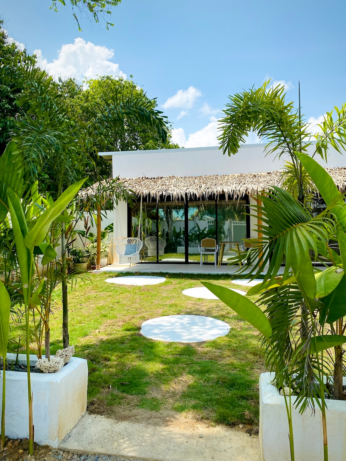 A modern structure is framed by lush greenery, showcasing a thatched roof and large glass doors. A manicured lawn is adorned with circular stepping stones, leading to a private terrace outfitted with outdoor seating. Tropical plants add a natural touch to the inviting entrance.