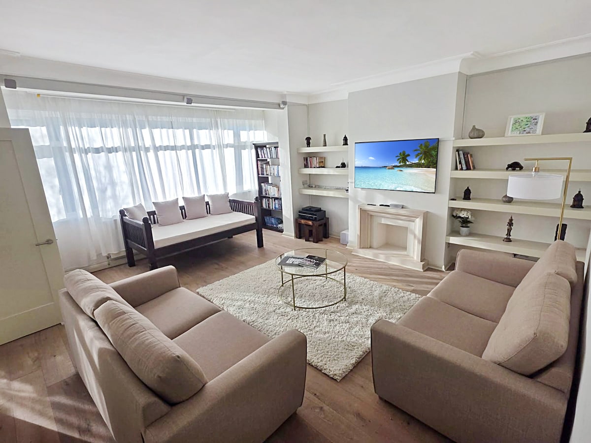 A bright living area features large windows draped with sheer curtains, allowing natural light to fill the space. Two comfortable sofas face a modern television mounted on a light-colored wall, while a round coffee table sits on a soft area rug, enhancing the relaxed atmosphere.
