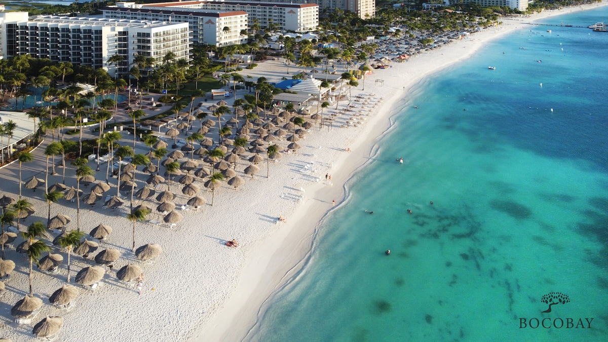 An aerial view captures a wide stretch of beach lined with thatched umbrellas and lounge chairs. The pristine white sand meets clear turquoise waters, while a series of beachfront accommodations are visible in the background, showcasing a vibrant coastal scene.