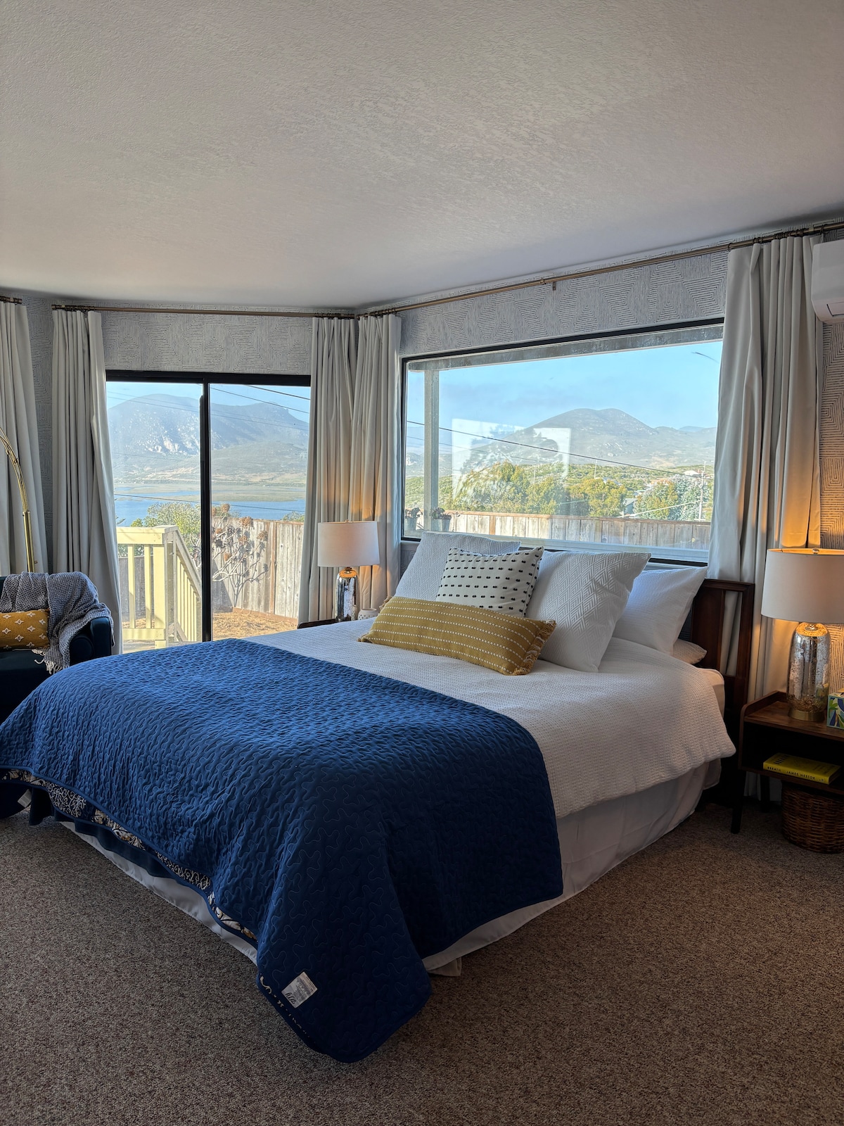 A cozy sleeping area features a spacious bed dressed in crisp white linens and a textured blue quilt. Large windows frame panoramic views of mountains and the surrounding landscape, while soft drapes add a calm touch. Ambient lighting is provided by bedside lamps on either side.
