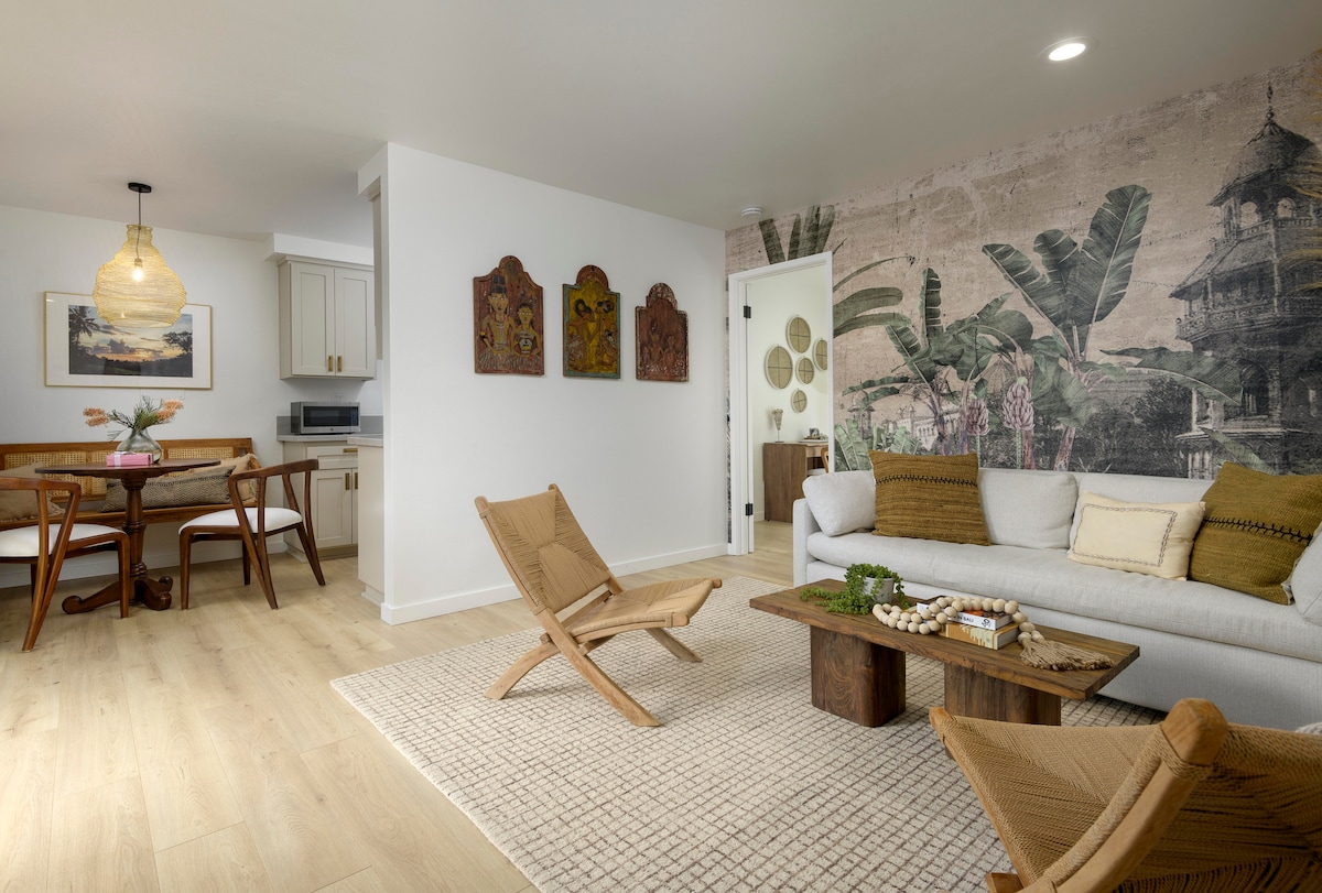 A welcoming living area features a modern sofa adorned with earthy tones and a textured coffee table. A mural depicting tropical foliage adds an exotic touch. Natural light illuminates the space, which is complemented by two wooden lounge chairs and a dining area in the background.
