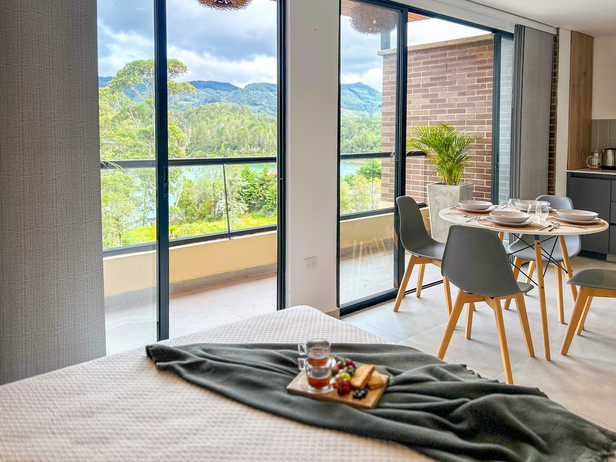 A spacious room features a comfortable bed, complemented by a tray of snacks and a drink. Large glass doors lead to a private balcony, providing an expansive view of the surrounding greenery and distant hills. A dining area with modern chairs is visible in the background.