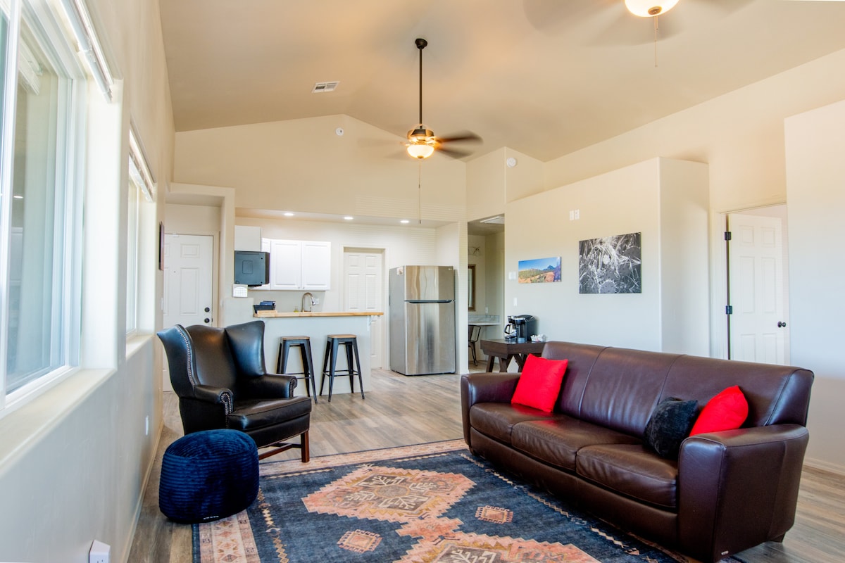 A spacious living area is highlighted by a dark brown leather sofa adorned with two red accent pillows. A dining area with a bar-style counter and high stools is visible in the background, along with a modern kitchen featuring stainless steel appliances. Large windows provide natural light.