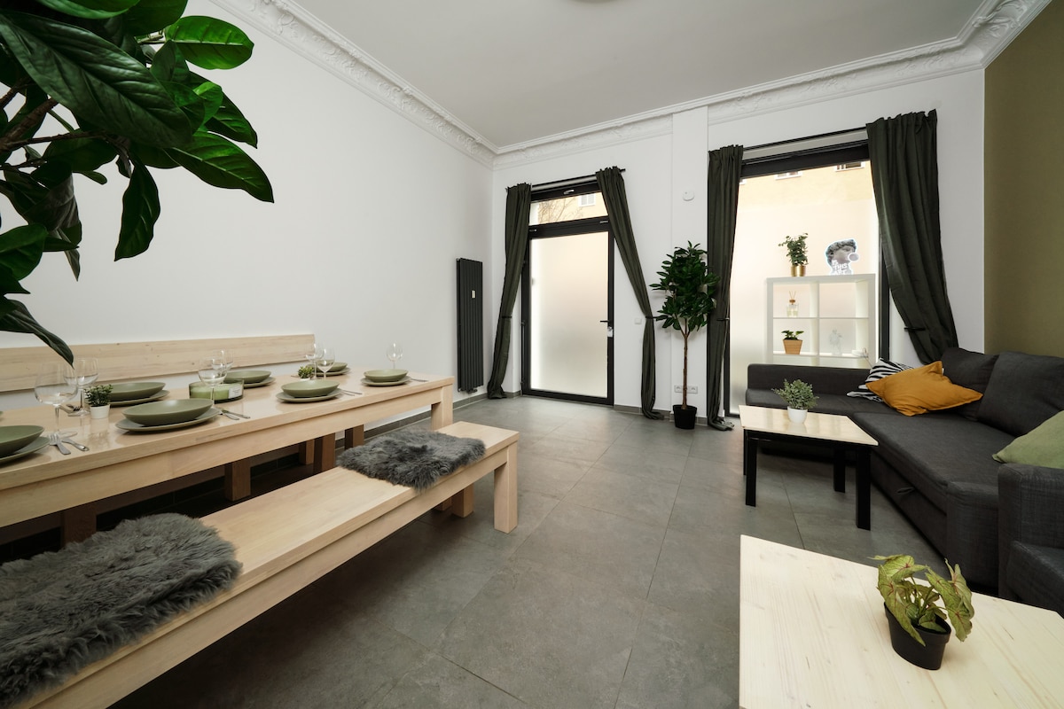 The living and dining area features a long wooden table set for meals, complemented by plush seating. Greenery is present with potted plants adding a touch of nature. Large windows with curtains allow natural light to illuminate the space, creating a calm atmosphere.