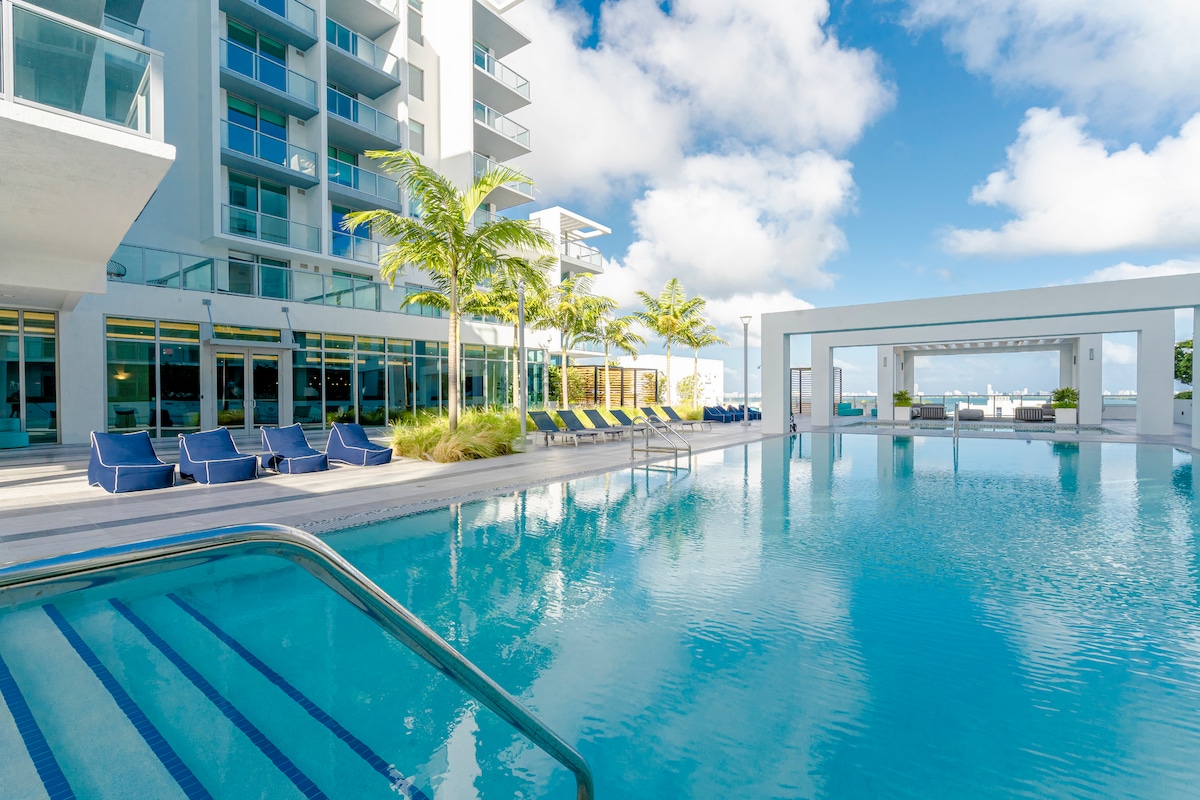 A resort-style pool area features a sparkling blue expanse of water, surrounded by lounge chairs and palm trees. A shaded cabana provides seating for relaxation. The clear sky and soft clouds create a bright, airy atmosphere above the pool deck, offering a serene outdoor space.