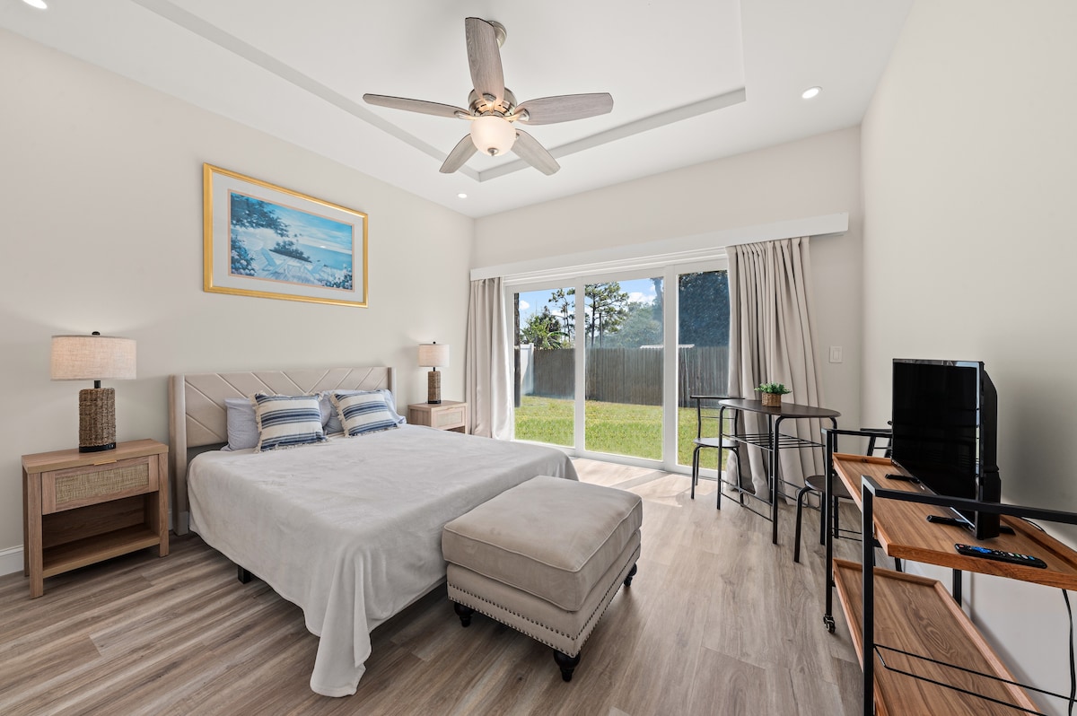 A generously sized sleeping area features a spacious bed, complemented by an ottoman and two nightstands with lamps. Natural light floods the room through sliding glass doors that open to a private outdoor space, enhancing a sense of tranquility and connection to nature.