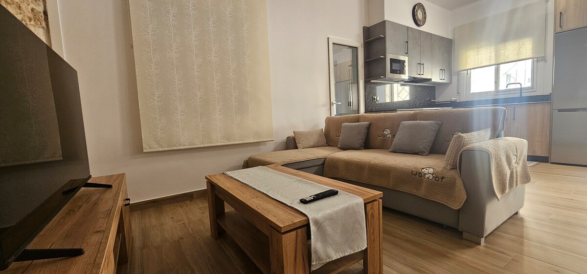 A modern living area features a comfortable sectional sofa adorned with light grey cushions and a soft throw blanket. A wooden coffee table sits in front, while a sleek flat-screen TV is positioned against the wall. Natural light enters through a nearby window, complemented by a patterned curtain.