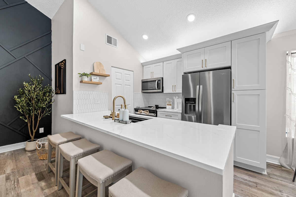A modern kitchen features light grey cabinetry and stainless steel appliances. A spacious white countertop with bar seating for four is highlighted. Natural light enters through a nearby window, illuminating the stylish design. A plant adds a touch of greenery to the decor.