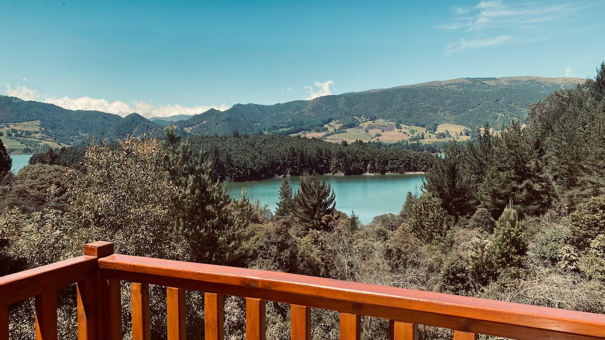 A wooden terrace railing frames a scenic view of a tranquil body of water surrounded by green hills and trees. The clear sky above enhances the natural beauty of the area, inviting peaceful contemplation and relaxation in the countryside.