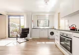 A bright, open kitchen and living area features sleek cabinetry and modern appliances. A large glass door offers views of the outdoor space, while a comfortable chair is positioned near the window, allowing natural light to fill the room. A washing machine is integrated into the cabinetry for convenience.