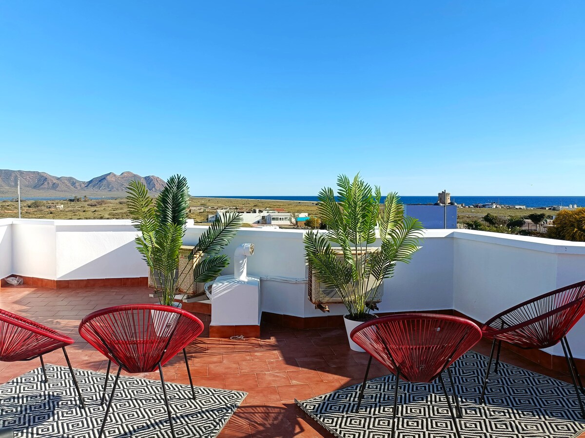 A shared rooftop terrace is depicted, featuring two red chairs positioned on a patterned rug. Lush green plants create a serene atmosphere, while expansive views of the blue sea and distant mountains serve as a backdrop, capturing the essence of the natural surroundings.
