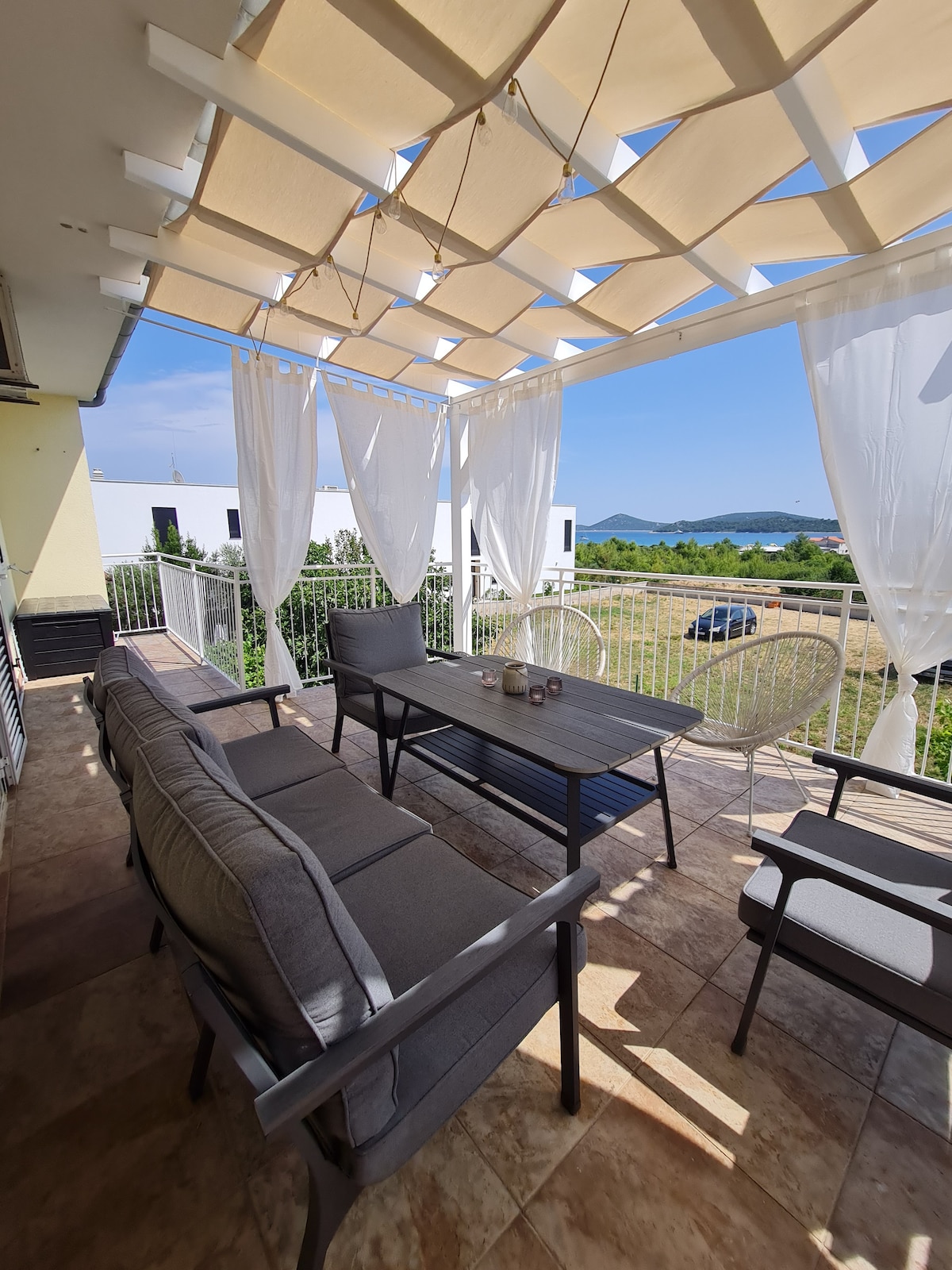 A spacious terrace features comfortable seating arrangements, complemented by a dining table. Light, airy curtains gently sway in the breeze, and a scenic view of the surrounding landscape and sea is visible in the background. The area is well-lit by natural sunlight.