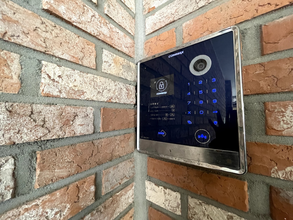 An electronic door entry system is mounted on a brick wall, featuring a keypad with illuminated numbers. The modern design incorporates a camera for added security, seamlessly integrated with the rustic texture of the exposed brick.