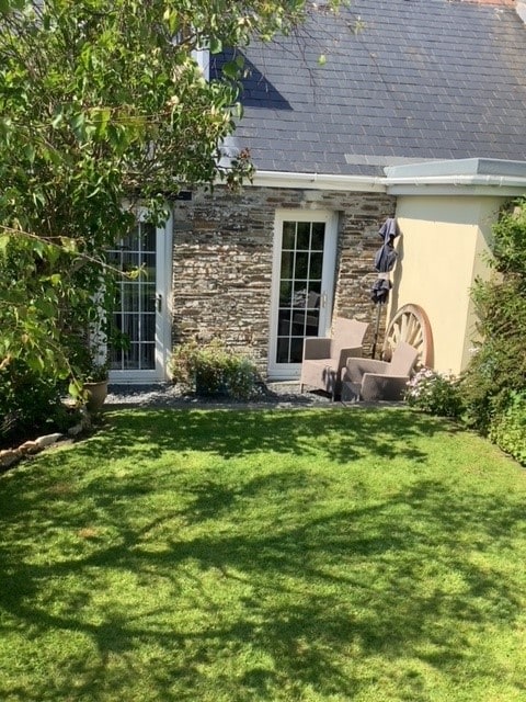 A well-maintained garden area is presented, featuring a neatly manicured lawn and stone walls. Two outdoor chairs are positioned near the entrance, surrounded by greenery and flowering plants. Sunlight enhances the inviting outdoor space, perfect for relaxing in a tranquil setting.