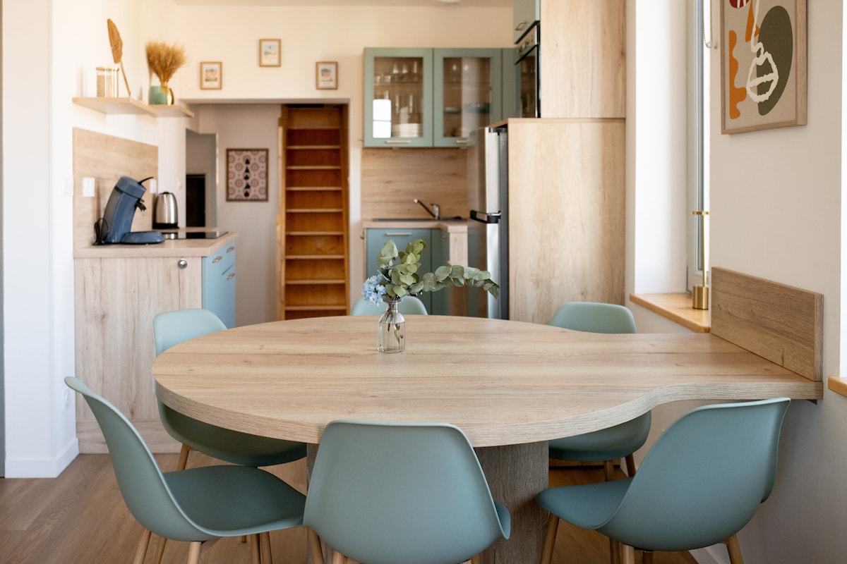A modern open living space features a round wooden dining table surrounded by six light blue chairs. A small vase with greenery is placed at the center of the table. The kitchen area is visible in the background, showcasing wood and glass cabinetry.