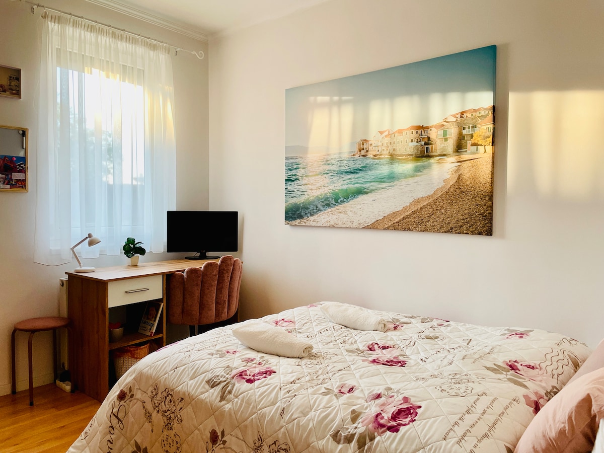 A bright sleeping area is presented, featuring a spacious bed adorned with a floral-patterned comforter. A desk with a monitor is positioned nearby, complemented by a pink upholstered chair. A large artwork depicting a coastal scene hangs above the bed, with natural light filtering through sheer curtains.