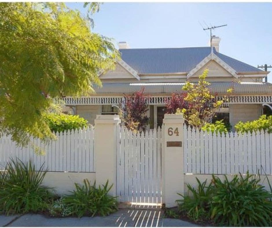 A charming heritage-listed home is showcased behind a low white picket fence. Lush greenery and flowering plants surround the entrance, creating an inviting appeal. The exterior features traditional architectural details, with a gabled roof and decorative wooden trims evident on the facade.