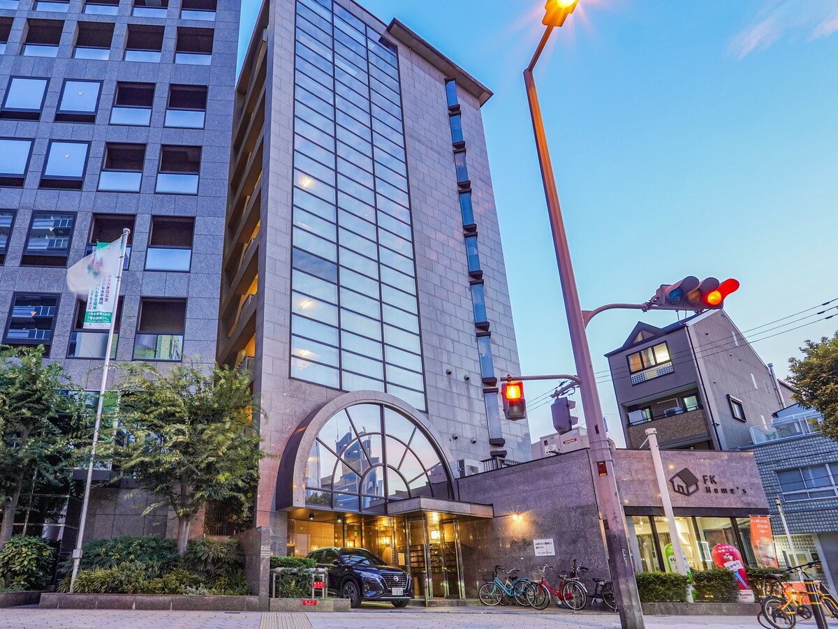 A modern apartment building is featured with a prominent glass façade, showcasing large windows that allow for natural light. The entrance is marked by a curved archway, surrounded by greenery and a pedestrian-friendly street. Bicycles are parked outside, adding to the urban setting.