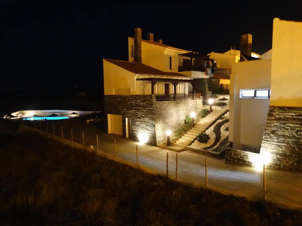 The exterior of a multi-level building is illuminated softly by landscape lighting at night. A pathway lined with stones leads to the entrance. In the background, a pool is visible, reflecting the night sky, surrounded by well-kept landscaping.