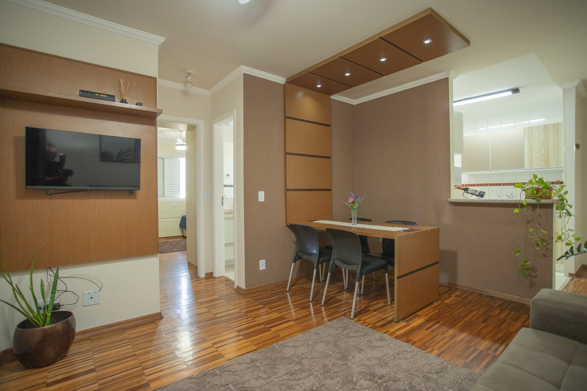 The living and dining area is depicted with a wooden floor, featuring a wall-mounted TV and a sleek dining table with four chairs. Natural light enhances the warm tones of the walls, complemented by decorative plants positioned in the corner.