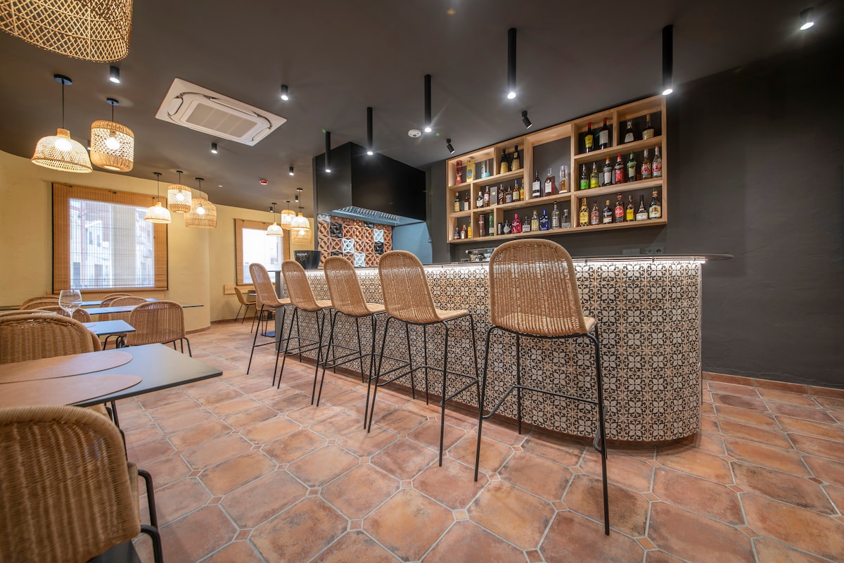 A modern bar area features a patterned counter with high stools arranged along one side. Well-lit shelves display an assortment of beverages. The warm ambiance is enhanced by decorative pendant lighting and tiled flooring, creating a welcoming space for guests.
