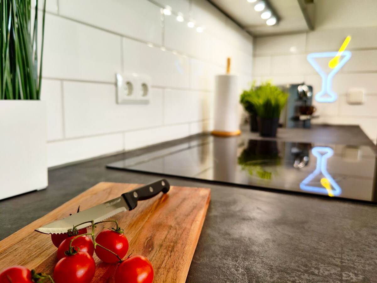 The kitchen features modern appliances and sleek countertops. A wooden cutting board displays fresh tomatoes next to a knife. Potted plants add a touch of greenery, while a neon cocktail sign reflects on the glossy surface, enhancing the kitchen's contemporary design.