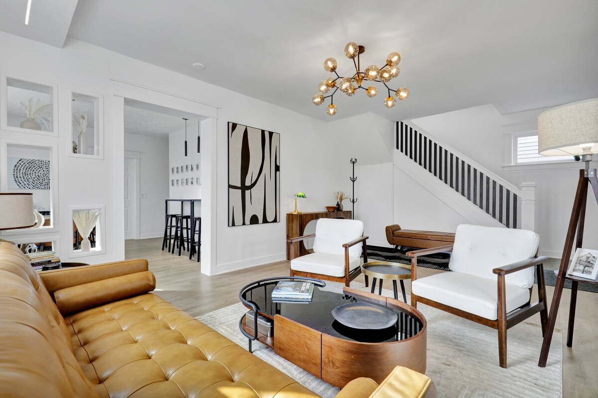 A modern living area features a stylish brown sofa paired with white armchairs. A sleek coffee table sits in the center, complemented by a unique light fixture above. The room is spacious, with accented artwork on the walls and an inviting staircase in the background.