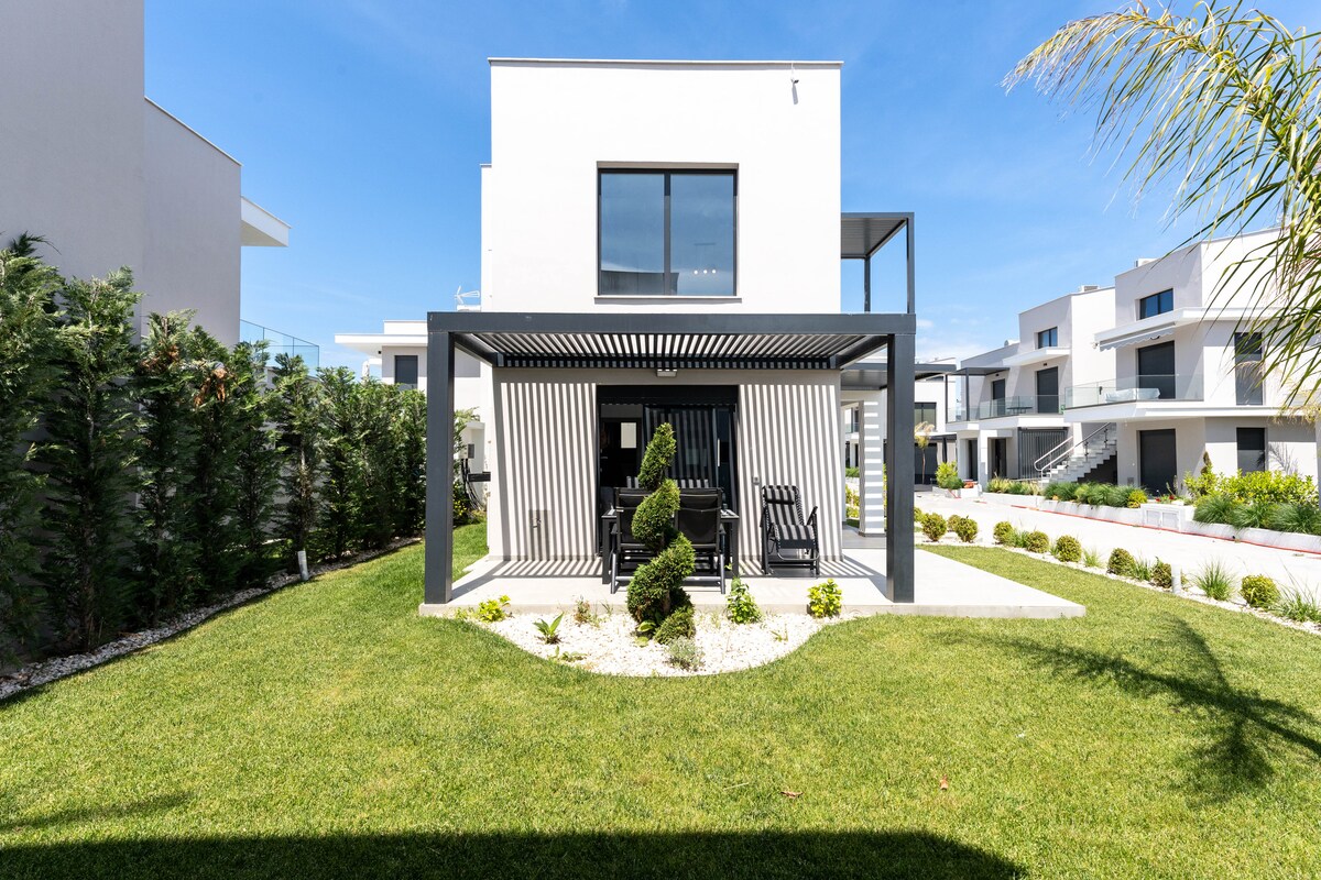 A modern outdoor space is showcased, featuring a shaded seating area beneath a stylish pergola. Neatly trimmed greenery and ornamental shrubs surround a manicured lawn, enhancing the contemporary architecture of the surrounding buildings.