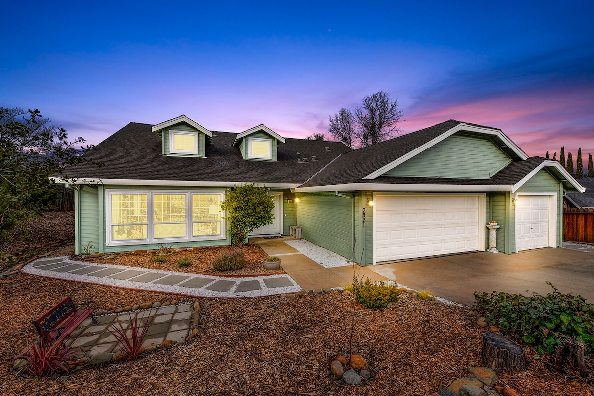 The exterior of a modern home is shown at dusk, with a soft purple sky in the background. The structure features a light green facade and multiple gabled roofs. The front yard is landscaped with low shrubs and stone pathways leading to a two-car garage.