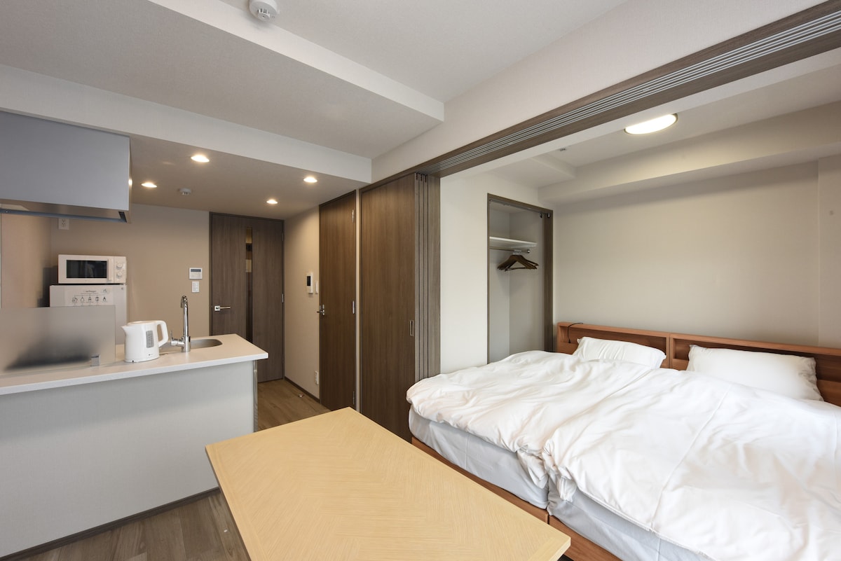 An open layout showcases a cozy sleeping area featuring two single beds with neatly arranged bedding. A wooden table sits in the foreground, while a kitchenette with modern appliances is visible beyond. Soft lighting contributes to a relaxed ambiance.