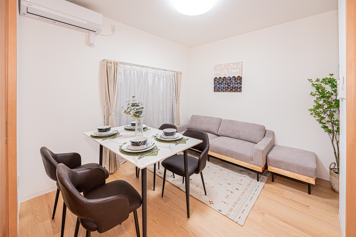 An open living space features a dining table set for four, with simple tableware placed atop a white tablecloth. A comfortable gray sofa and an accent chair are present, alongside a potted plant. Soft natural light enters through a window covered by sheer curtains.