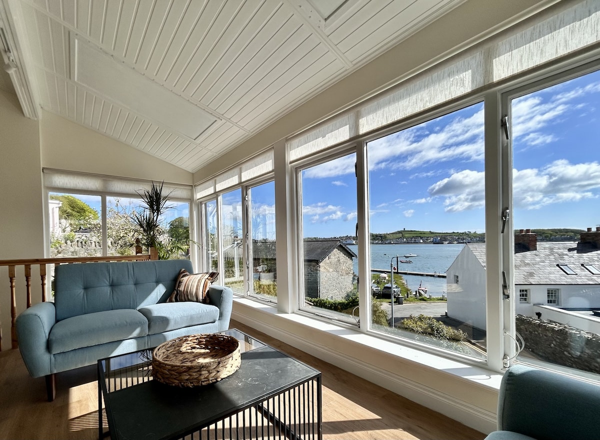 A sunlit sunroom is featured, showcasing expansive windows that provide views of the quay and nearby buildings. A light blue couch is positioned next to a coffee table, with decorative elements enhancing the serene atmosphere. The bright surroundings are complemented by clear skies and a view of the water.